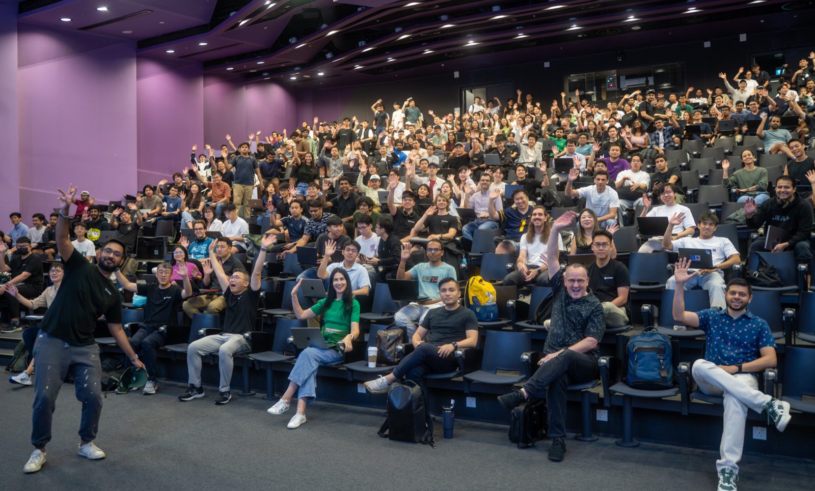 A packed lecture theatre of builders with laptops, waving at the camera during a hands-on AI workshop